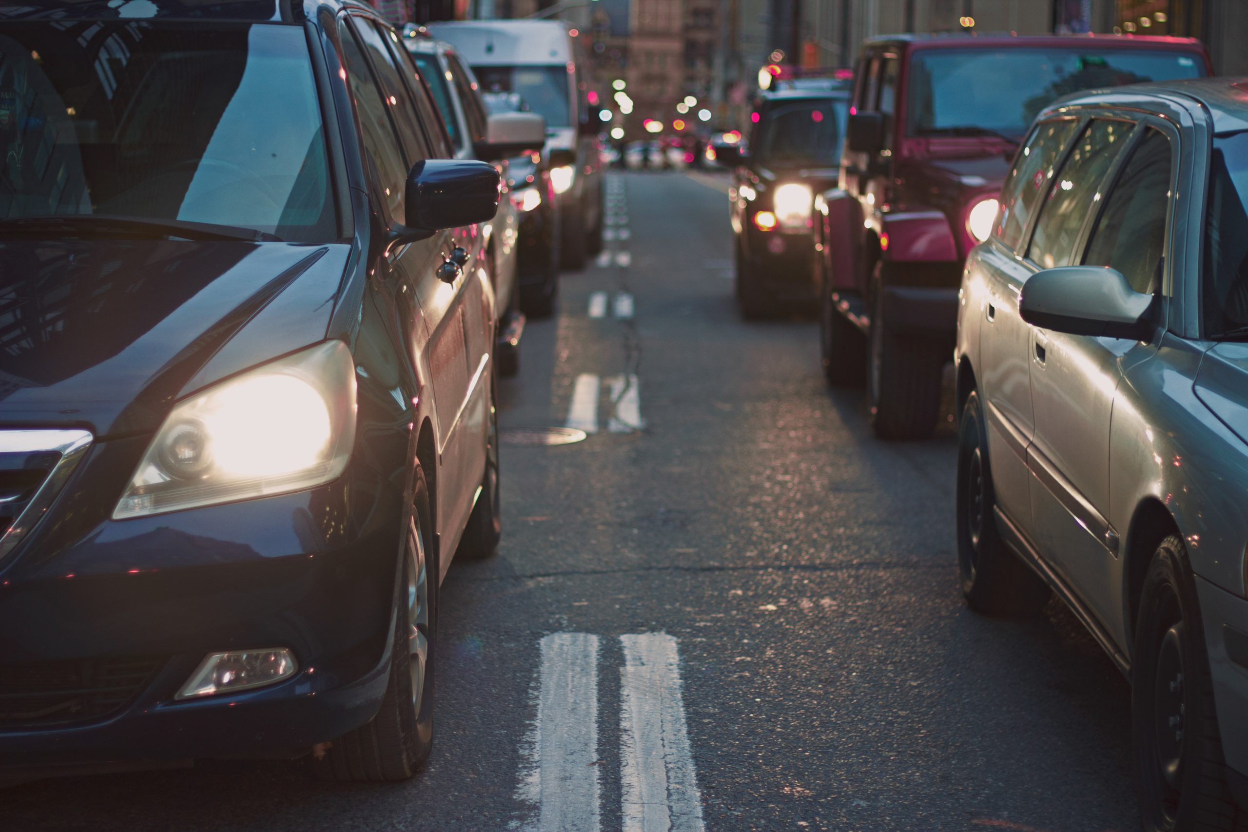 A busy urban street is filled with vehicles in both directions, including cars and vans. Headlights are on as dusk falls, and buildings line the street. The atmosphere suggests evening traffic congestion, where tailgating can amplify safety concerns amid the rush.