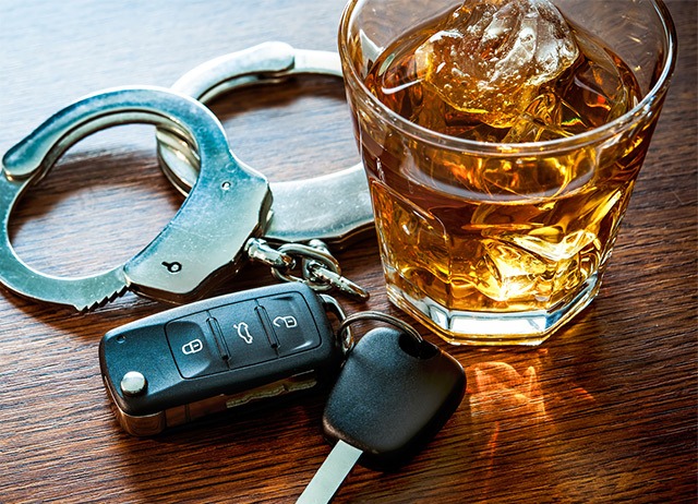A glass of whiskey with ice sits on a wooden table next to a pair of handcuffs and a set of car keys.