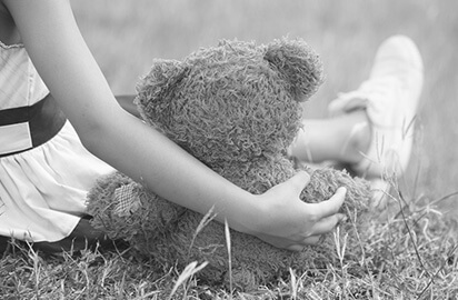 A child sitting on grass holds a teddy bear. The child is wearing a dress and white sneakers. The scene is in black and white, creating a nostalgic and calm atmosphere.