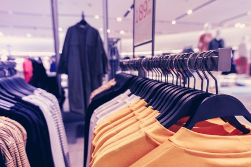 A clothing store display with various shirts on hangers. The foreground features yellow shirts, while darker clothing items are visible on hangers in the background. The setting is brightly lit with a modern design.