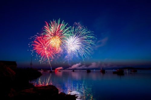 A vibrant fireworks display lights up the Texas night sky with red, green, and blue bursts. Reflections shimmer on the calm water below. Boats are scattered across the water, and rocky shores frame the scene under a deep blue evening sky—a perfect moment to ponder firework laws.