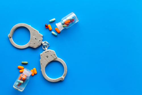 Handcuffs and small bottles of colorful pills and capsules are spread across a bright blue background, evoking the complex issues of drug possession in Texas or addiction. The image subtly hints at the legal defenses that might unfold in such scenarios.