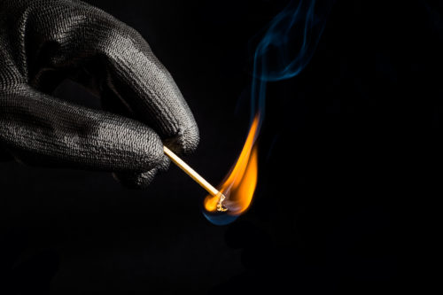 A gloved hand holds a lit match against a dark background, the small flame casting an eerie glow as wisps of blue smoke rise from the matchhead—capturing a moment that echoes the cautionary tales of Texas arson laws.