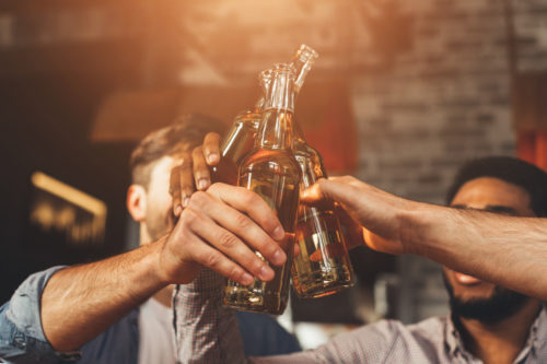A group of people clinking beer bottles in a celebratory gesture, with warm atmospheric lighting enveloping them. The background is blurred, spotlighting the raised bottles and the social gathering theme—a quintessential Texas toast, mindful of public intoxication laws.