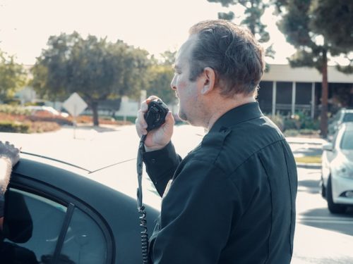 A police officer stands beside a car at a sobriety checkpoint, holding a radio microphone to his mouth. He's in uniform, and the scene unfolds outdoors in a sunny parking lot, with trees and buildings in the background.