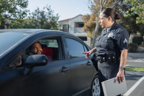 A police officer stands beside a black car, discussing DWI in Texas with the driver. The driver looks up at the officer, perhaps wondering when their license will be reinstated. Trees and houses provide a serene backdrop to this serious roadside conversation.