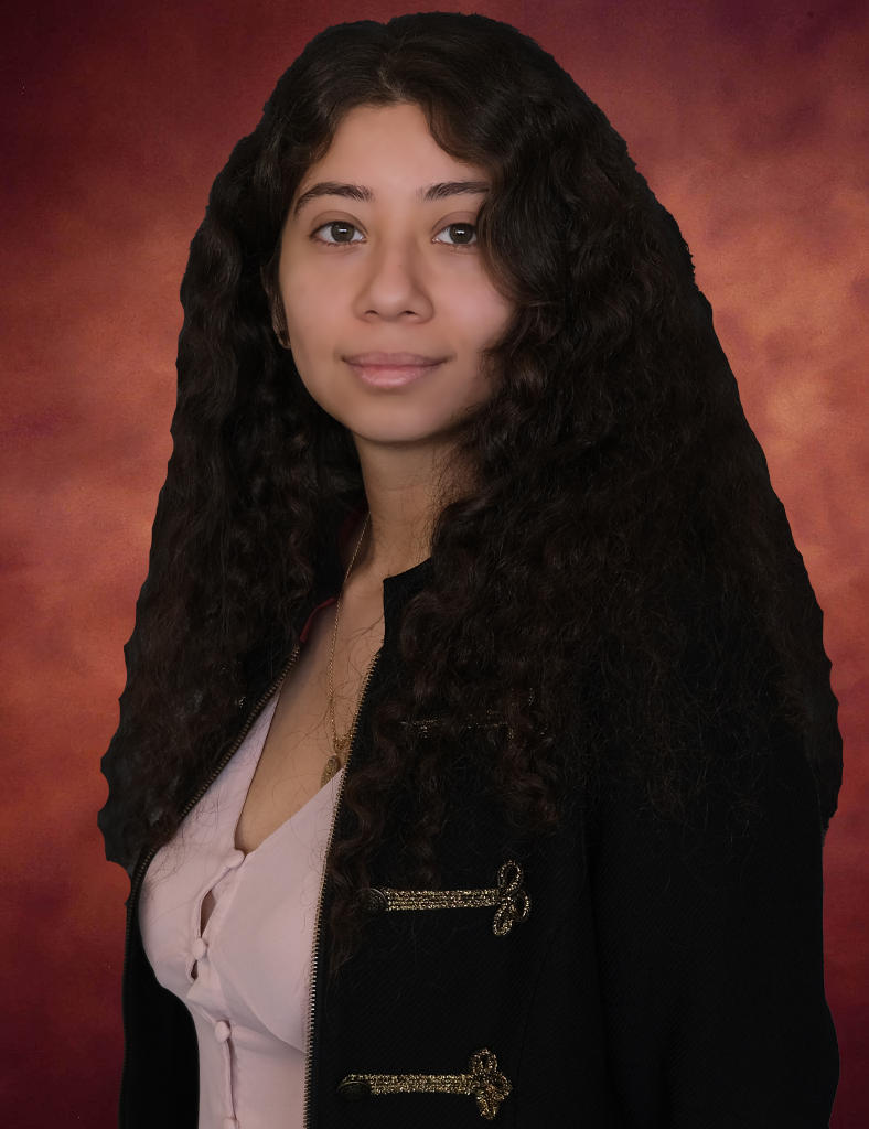 Rosa Palencia, with long, curly dark hair, poses against a warm, reddish-brown backdrop. She wears a black jacket with gold details over a light pink shirt. Her calm expression is captivating as she gazes slightly off-camera.