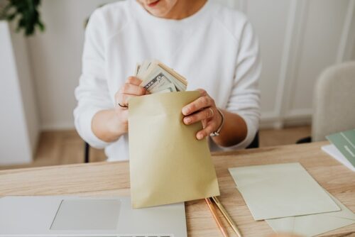 A person sits at a desk in Texas, discreetly slipping stacks of cash into a brown envelope. A laptop is open on the table nearby, surrounded by more envelopes and stationery items, hinting at potential bribery despite looming penalties.