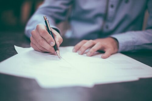 A person wearing a blue shirt is meticulously writing notes on a stack of white papers with a black pen, possibly preparing for a DWI case. The focus is on their hands, emphasizing concentration and dedication to the impending jury trial; the background is slightly blurred to maintain this focus.