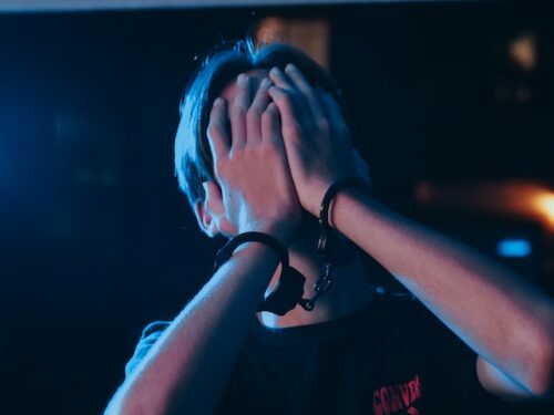 Under duress, a person in handcuffs shields their face with both hands, standing under dim, blue-tinted lighting. They are wearing a black shirt featuring a red logo on the chest. The dark background is blurred, casting an aura of mystery over the scene.