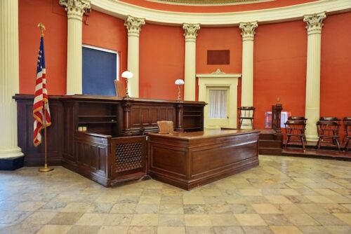 An empty courtroom with wooden desks and chairs, featuring tall white pillars, a U.S. flag, and echoes of Texas law. The room has a red wall and a large, ornate ceiling, reminiscent of the solemnity found in moments of jury duty and the weighty consequences that follow.