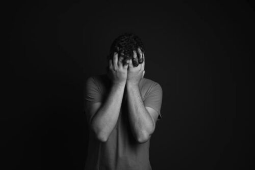 In a black and white photo, a person stands against a dark background, hands covering their face in anguish. Wearing a short-sleeved shirt, they embody the emotional struggle often seen at the intersection of mental health issues and Texas criminal cases.