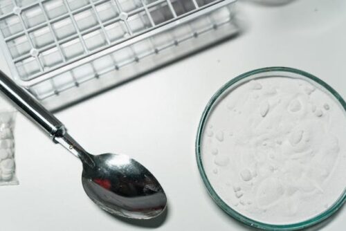 Close-up of a spoon next to a Petri dish containing a white powdery substance, reminiscent of the strict Texas cocaine laws, with a white grid structure partially visible in the background on a white surface.