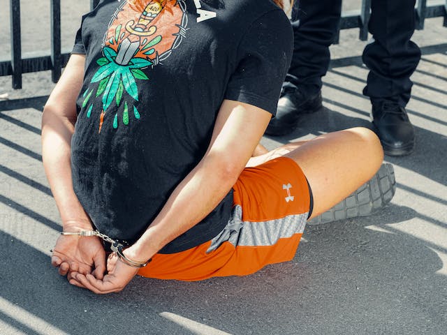 A person wearing an illustrated black t-shirt and orange shorts sits on the ground with hands cuffed behind their back, near a metal fence, while another person in dark clothing stands nearby.