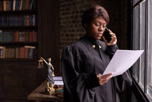 A person in a judge's robe stands by a window, holding a search warrant and talking on the phone. In the background are bookshelves and a Lady Justice statue.