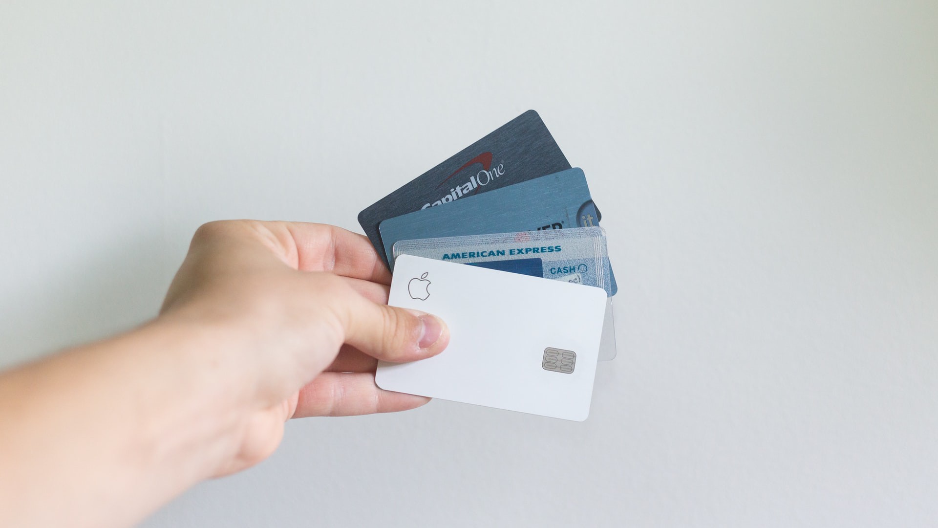 A hand holding four credit cards, fanned out. The front card is a white Apple Card, with American Express, CapitalOne, and Chase cards partially visible behind it. The background is plain and light-colored.