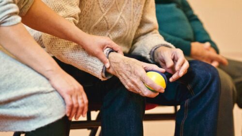 A caregiver gently supports an elderly person's arm as they hold a colorful stress ball, ensuring their safety and well-being. Another person sits nearby. The scene conveys care and support in a warm, comfortable setting, countering any fears of elder abuse with genuine compassion.