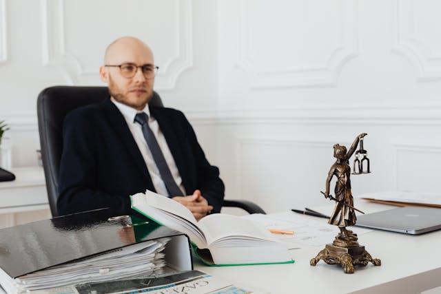 A man in a suit sits at a desk with open books, folders, and a Lady Justice statue in the foreground, suggesting a legal or office setting where he may be preparing as a character witness.