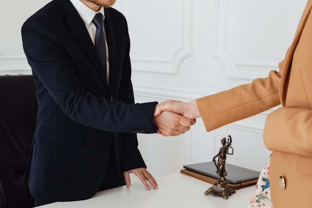 Two people in business attire shaking hands across a desk in an office, suggesting a meeting with a Criminal Defense Attorney. Only their torsos and hands are visible, alongside a small statue, folders, and documents on the desk.