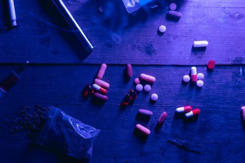 A wooden surface under purple lighting displays scattered pills in various shapes and colors, hinting at the shadows of drug possession. Nearby, a syringe lies ominously next to a small transparent bag with unknown contents.