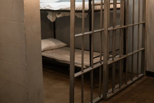 A dimly lit space reveals a small jail cell with metal bars and two bunk beds, each with white sheets and a pillow. The concrete floor and bare walls intensify the stark, confined atmosphere—a place to avoid if you wish to keep your freedom.