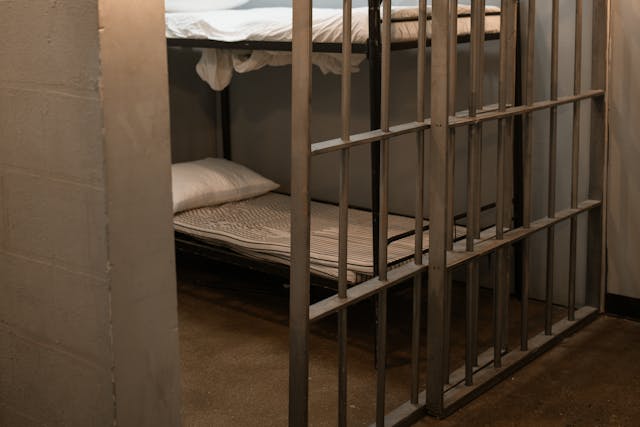 A jail cell with metal bars contains a bunk bed with white sheets and pillows, set against plain concrete walls on a bare floor—a stark reminder of why it’s best to avoid jail.