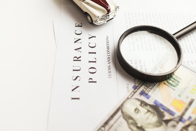 A magnifying glass, a toy car, and a $100 bill rest on top of an insurance policy document, highlighting the details that can affect your car insurance rates.