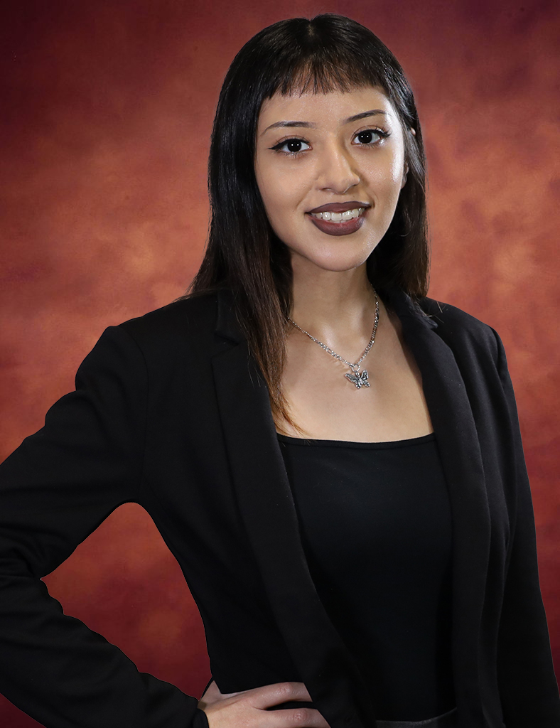 Italimi Nunez, a woman with long dark hair and short bangs, wears a black blazer and top, smiling at the camera. She elegantly sports a necklace with a pendant. The background is a warm, reddish-brown gradient.