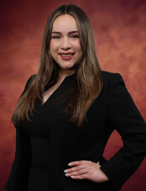 Karina Ochoa, with her long brown hair, smiles confidently as she stands with a hand on her hip, wearing a sleek black blazer against a warm, rust-colored background.