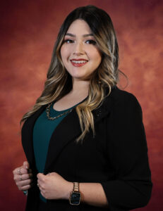 Liliana Salas, with her long, wavy hair, smiles warmly in a teal shirt and black blazer. Her ensemble is beautifully accented by a gold necklace and watch against a warm, gradient brown backdrop.