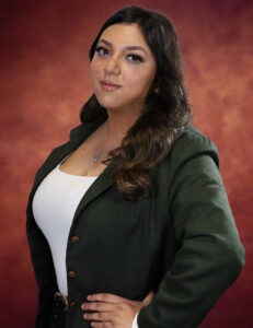 Nashelly Salazar, with long dark hair, poses confidently against a warm, reddish-brown background. Wearing a dark green blazer over a white top, her expression is calm and self-assured.