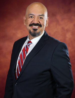 Ruben Anthony Nájera, a bald man with a mustache and goatee, stands in front of a warm, brownish-red background. Dressed in a dark suit, white shirt, and red-striped tie, he smiles confidently at the camera.