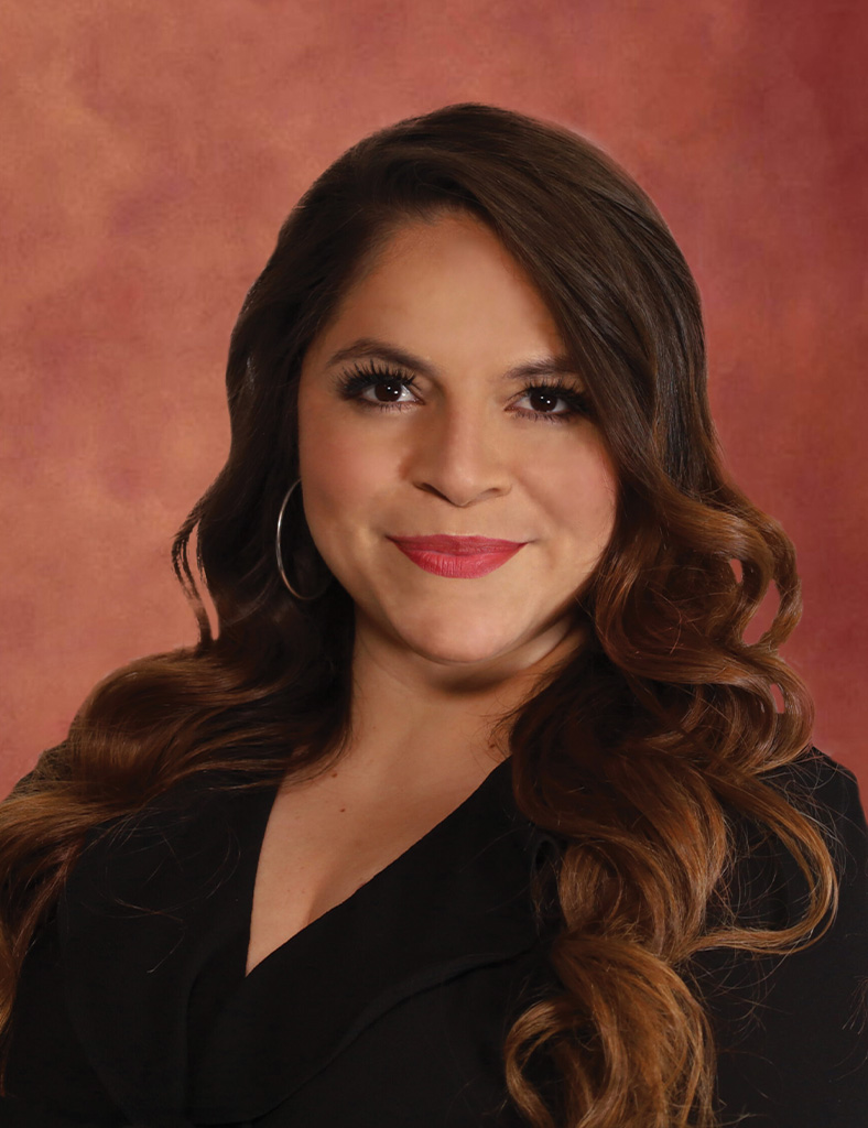 Fany Broussard, a woman with long, wavy brown hair and hoop earrings, smiles softly. She is wearing a black outfit against a warm, reddish-brown background. Her makeup is natural, with a focus on the eyes and lips, enhancing her graceful presence.