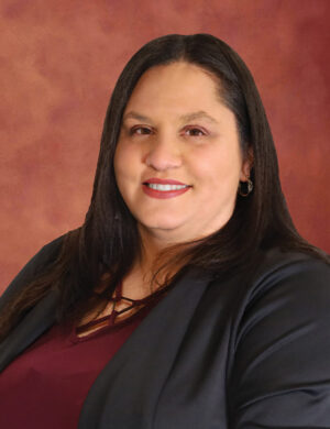 Maria Luisa Gonzalez, a woman with long dark hair, is wearing a black blazer and burgundy top, smiling warmly against a maroon background.