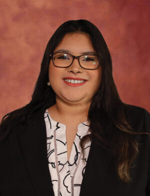 Virginia Zubia, a woman with long dark hair and glasses, smiles at the camera. She is wearing a black blazer over a white patterned blouse. The background is a warm, muted pinkish-brown color.