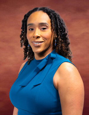 Yvonne Olusi-Ogadi, with her shoulder-length curly hair, is wearing a sleeveless blue dress. Against a warm brown background, she gazes at the camera with a slight smile.