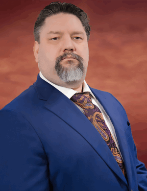 Bearded Davis Taylor stands confidently against a reddish-brown background, donning a blue suit and paisley tie, looking directly at the camera.