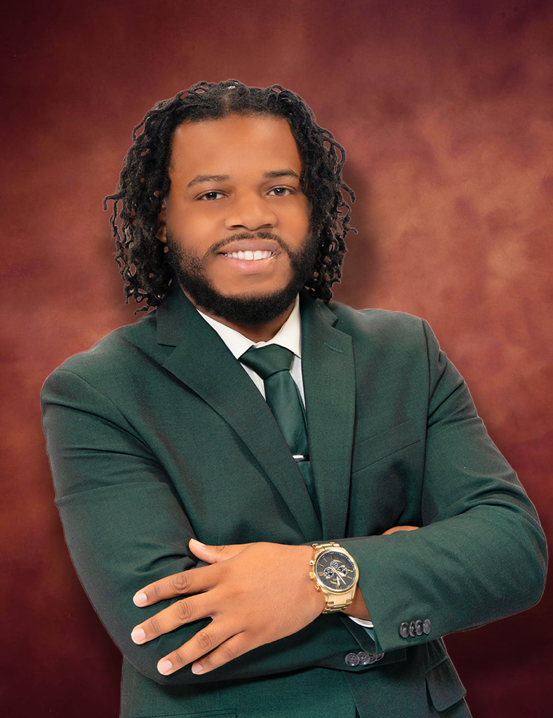 Dominick Dunlap, a man with curly hair, is smiling in his dark green suit paired with a white shirt and green tie. With arms confidently crossed, he sports a gold watch against the warm brown backdrop.
