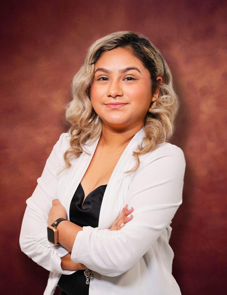 Jackie Lucio, with curly, shoulder-length light hair, is wearing a white blazer over a black top. They are smiling gently, with arms crossed against a maroon-brown textured background.