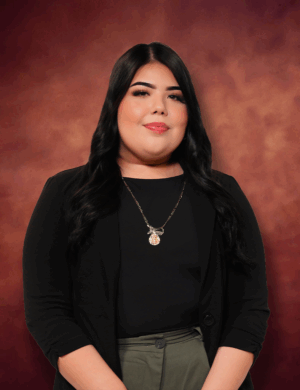 A person with long dark hair wearing a black blazer and green pants stands against a warm, blurred background. They are smiling gently and wearing a pendant necklace.