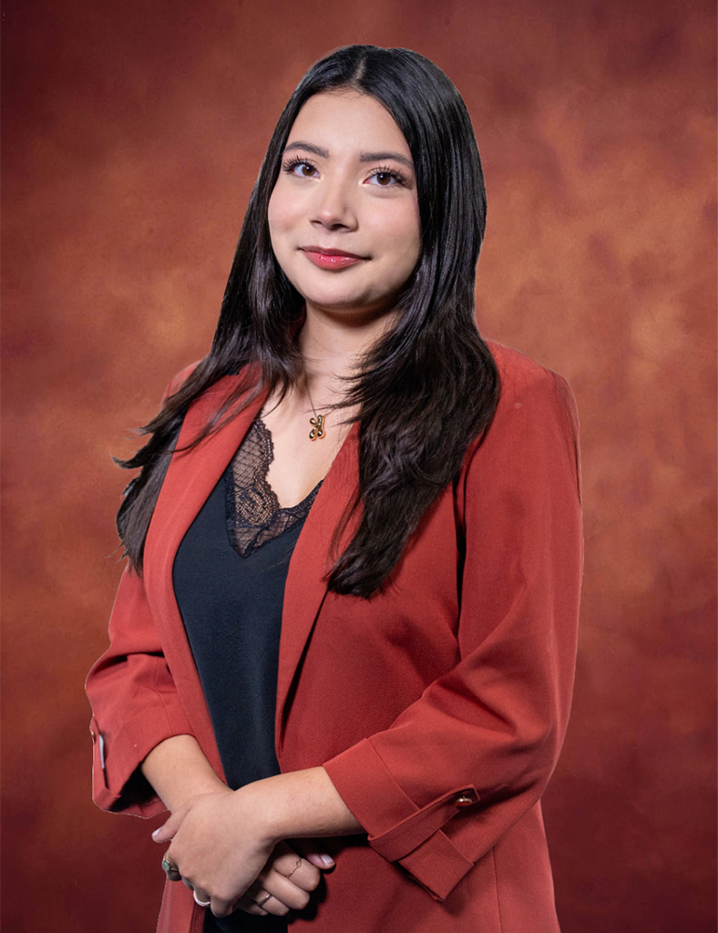 Katelyn Rocha, a woman with long dark hair, stands against a warm, mottled brown background. She is wearing a rust-colored blazer over a black top, looking slightly to the side with a calm expression.