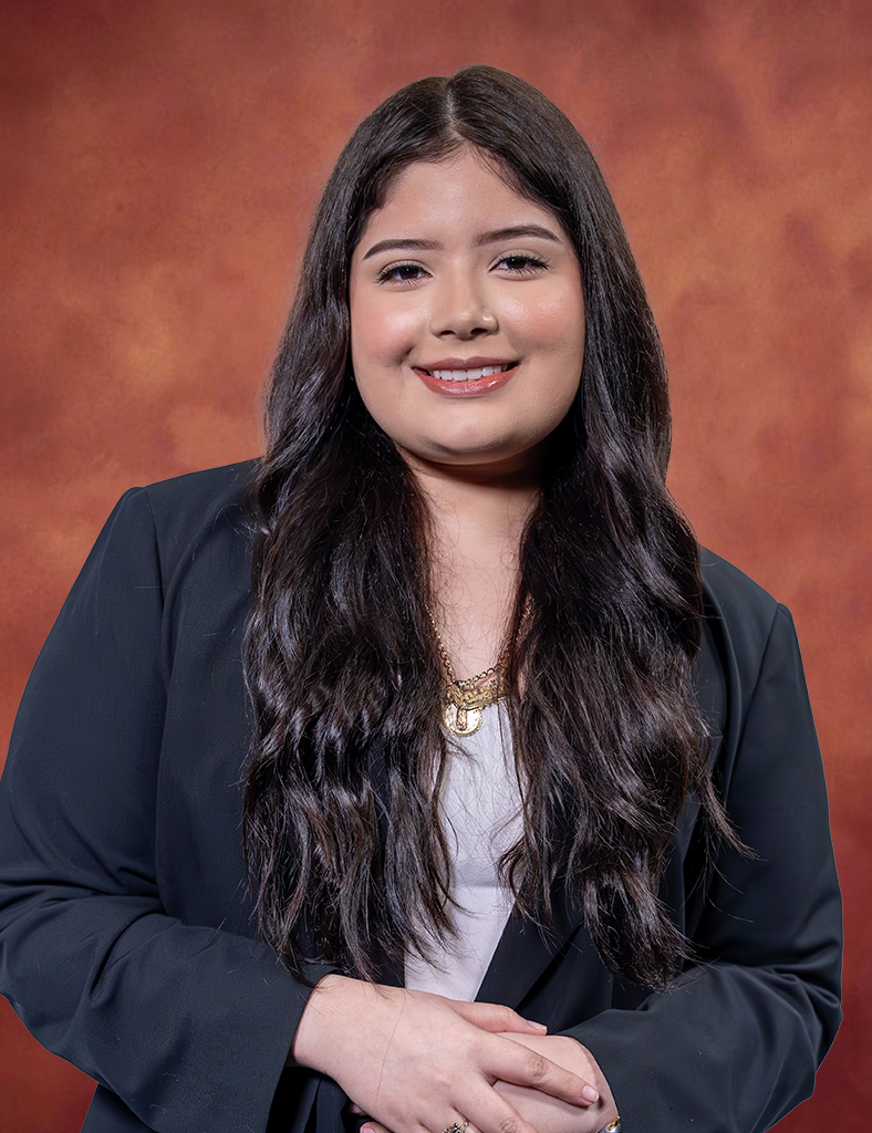 Katelyn Rocha, with long, wavy hair, smiles while wearing a dark blazer over a light top. She stands against a warm, textured brown background.