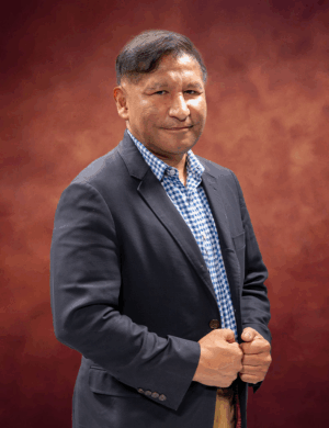 A man in a dark blazer and blue checkered shirt stands against a warm, brownish-red background. He has short hair and is smiling slightly, with hands grasping the sides of his blazer.
