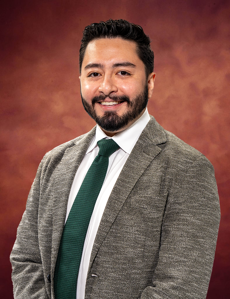 Jose Carrera, a man with short dark hair and a beard, is smiling warmly. He dons a gray suit jacket, white shirt, and green tie against a backdrop of warm brown tones.