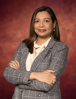 Alejandra Fiedler stands confidently with arms crossed, her long hair cascading over a gray checkered blazer paired with a light blouse. The warm, reddish hue in the background complements her poised presence.