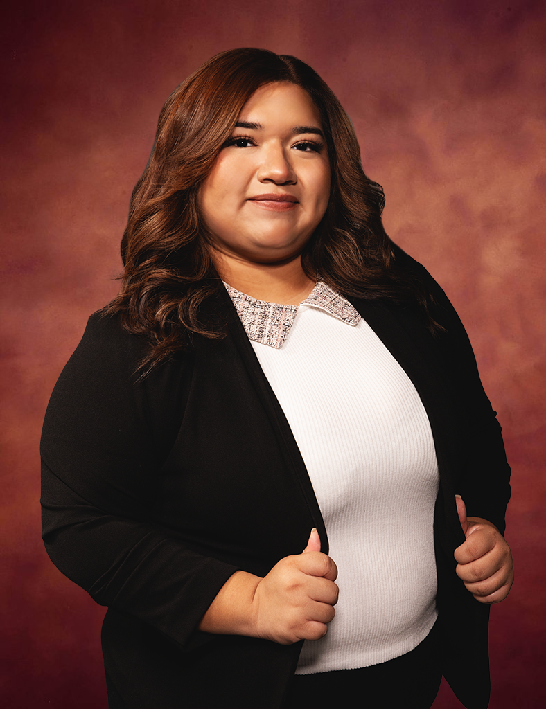Jackie Lucio, with her long brown hair and a radiant smile, stands against a warm-toned backdrop. She elegantly holds the lapels of her black blazer, worn over a white top with a sparkling collar.