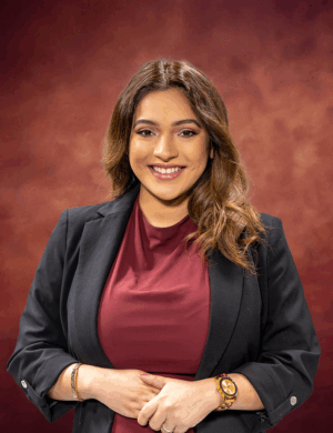 A woman with long, wavy hair, resembling Karyme Olmos, smiles while wearing a black blazer over a maroon top. The background is a warm brownish-red gradient. She wears a gold watch and bracelets, and her hands are gently clasped in front of her.