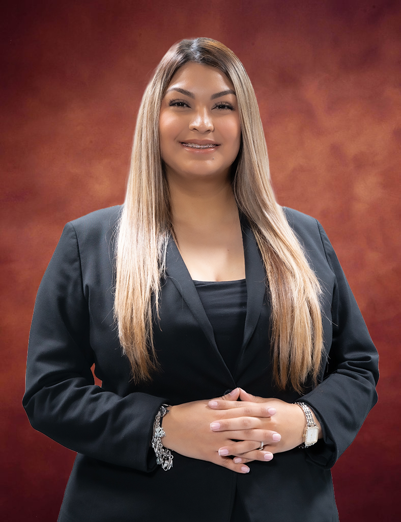 Kassandra Gonzalez, with her long, straight hair, smiles confidently against a warm, textured background. She wears a dark blazer and has her hands clasped in front.