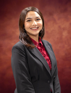 Vanessa Ruiz, a woman with brown hair, smiles while wearing a black blazer over a red blouse. She is posed in front of a warm-toned, textured background.
