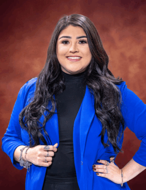 Yarezith Salazar, a woman with long dark hair, stands confidently against a warm, brownish-red backdrop. She wears a bright blue blazer over a black top, accessorized with rings and bracelets, and smiles warmly at the camera.